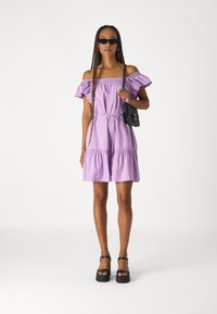 Robe lilas décolletée avec des manches volantées, taille cintrée et jupe à volants. Associée à des chaussures à plateforme noires et un petit sac noir.