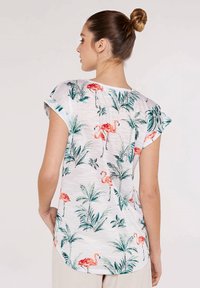 Haut blanc à manches courtes avec un motif de flamant rose et de feuilles tropicales. Tissu doux et texturé avec des manches retroussées et un ourlet arrondi.