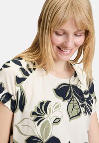 Kwiatowy t-shirt w kolorze kremowym, z ciemnoniebieskimi i zielonymi wzorami liści. Miękka tkanina z okrągłym dekoltem i rękawami na ramiączkach, zdobiona dekoracyjnymi detalami.