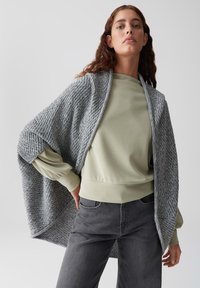 Grau gestrickter Schal über einem hellgrünem Sweatshirt, das einen lockeren Schnitt, lange Ärmel und ein strukturiertes Muster aufweist. Kombiniert mit dunkelgrauen Hosen.