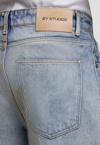 Artimas šviesiai mėlyno džinsinio audinio kelnių galinės kišenės vaizdas su šviesiai rudos odos ženklu, pažymėtu "2Y STUDIOS".