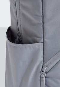 Gray fabric backpack featuring a front zip pocket and a side mesh pocket. Textured material with visible stitching and zip closure details.