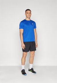 Camiseta atlética azul de manga corta, pantalones cortos negros, calcetines blancos y zapatillas negras con acentos verdes. El modelo está de pie de frente sobre un fondo liso.