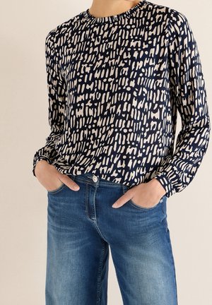 Persoon draagt een blouse met lange mouwen in marineblauw en wit patroon en blauwe jeans met handen in de zakken tegen een neutrale achtergrond.
