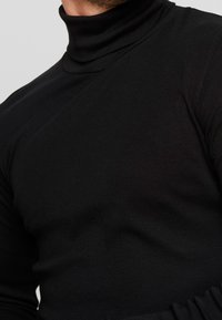 Homme portant un pull à col roulé noir ajusté, coupé au-dessus du menton et à la taille, sur un fond clair uni.