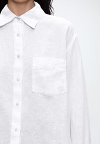 Chemise blanche en lin boutonnée avec col pointu, poche poitrine côté gauche, portée par une personne aux cheveux bouclés foncés sur un fond clair.