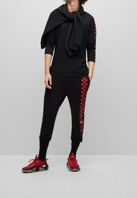 Czarna koszula z długim rękawem z żebrowanymi rękawami, w połączeniu z czarnymi dresami w czerwono-szare kratki. Czerwone sneakersy z czarnymi akcentami.