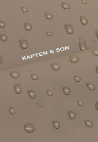 Wasserabweisender Stoff in einem gedämpften Braunton, versehen mit mehreren Wassertropfen. Bedruckter Text "KAPTEN & SON" in Weiß. Glatte Textur.
