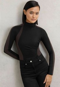 Top negru cu mâneci lungi și guler fals; are panouri laterale de un maro închis și o siluetă ajustată, asociat cu blugi negri cu talie înaltă.
