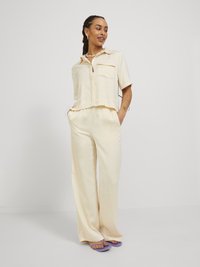 Camicia color beige chiaro, a maniche corte, con colletto, chiusura con bottoni e due tasche sul petto; abbinata a pantaloni beige larghi e coordinati.