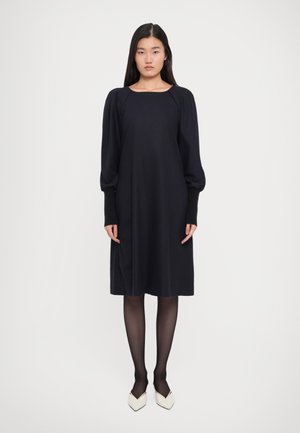 Woman standing wearing a black knee-length dress with puffed long sleeves, black sheer tights, and white pointed-toe heels against a white background.