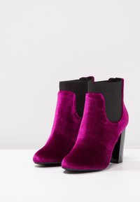 Botas de tobillo de terciopelo fucsia con paneles laterales elásticos negros y un tacón grueso negro. Textura suave y silueta moderna y elegante.