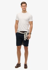 Pánský outfit se skládá z bílé košile, námořnických kraťasů, šedého svetru uvázaného v pase, hnědých kožených bot na lodě a hnědého opasku.