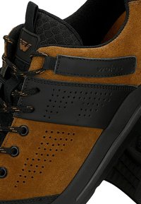 Sneaker in suede marrone e pelle nera con una tomaia texture, chiusura con lacci e dettagli laterali perforati. Nome del marchio "wojas" visibile.