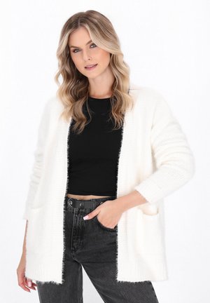 Weißer Strickcardigan mit offenem Front, strukturiertem Finish, langen Ärmeln und seitlichen Taschen, kombiniert mit einem schwarzen Oberteil und dunklen Jeans.