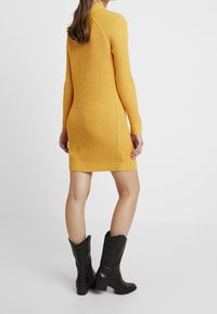 Strickkleid in leuchtendem Gelb mit taillierter Passform, langen Ärmeln, geripptem Stoff und Mid-Thigh-Länge. Kombiniert mit schwarzen Cowboy-Stiefeln.