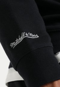 Manica nera in tessuto con il logo ricamato bianco "Mitchell & Ness" vicino al polsino a coste, indossata sopra uno strato bianco.