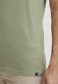 Oliva zöld pamut póló kerek nyakkivágással, rövid ujjakkal és kis márkacímkével a szegély bal oldalán. Sima textúra.