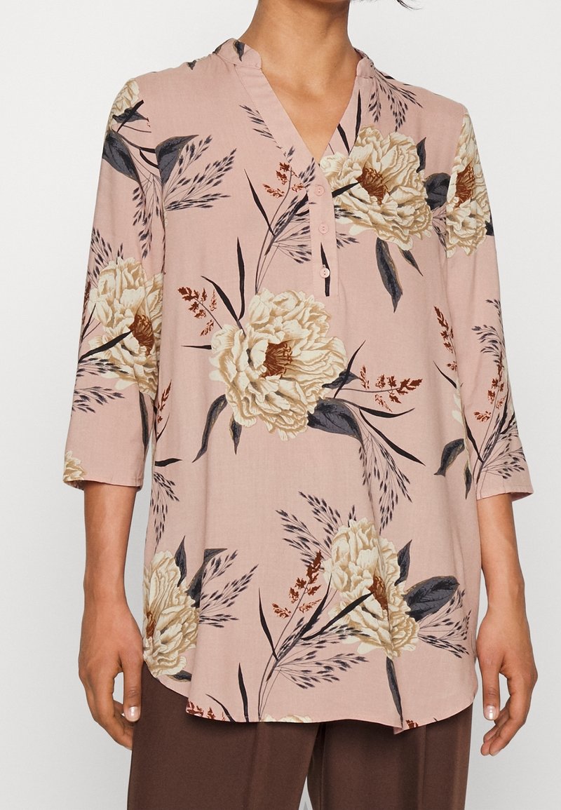 Blouse rose clair avec grands motifs floraux crème et marron, manches trois-quarts et col en V partiellement boutonné.