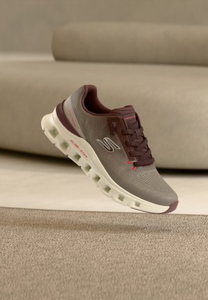 Barna és szürke Skechers tornacipő fehér talppal és piros díszítéssel, bézs szőnyeg felett lebegve egy modern szobában.