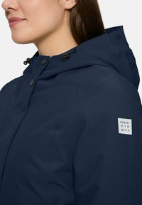 Woman wearing a navy blue hooded jacket with black buttons and a New View logo patch on the sleeve, shown from shoulder up.