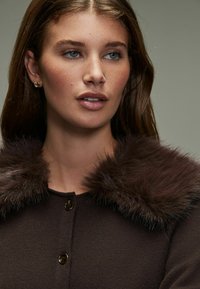 Bruine gebreide cardigan met een nepbont kraag, voorzien van zwarte knopen en een zachte textuur. De huid van het model heeft een natuurlijke finish, met neutrale make-up.