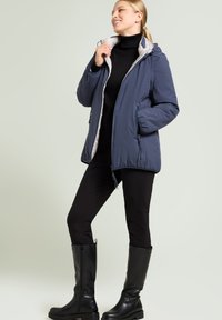 Marineblaue isolierte Jacke mit Reißverschluss vorne, Kapuze und Seitentaschen, kombiniert mit schwarzen, eng anliegenden Hosen und kniehohen schwarzen Stiefeln.