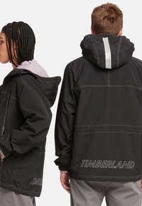 Chaqueta impermeable negra con costuras grises, capucha ajustable y detalles de franjas reflectantes. Presenta un logotipo de "TIMBERLAND" en la parte posterior.