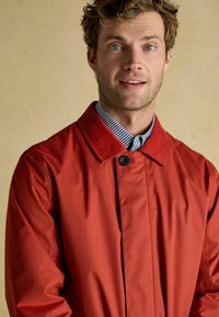 Raudona neperšlampama striukė su veliūro apykakle, vieno mygtuko užsegimu ir lygiu paviršiumi, vilkima per mėlyną ir baltą dryžuotą marškinėlį.