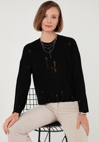 Črna pletena pulover z obrabljenimi detajli, okroglo dekolte, dolgi rokavi in ohlapen kroj, v kombinaciji s svetlo bež hlačami.