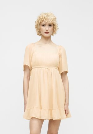 Young person with short curly blonde hair wearing a short, peach-colored, flowy dress with sheer sleeves against a white background.