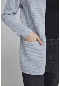 Ljusblå texturerad kofta med löst sittande passform, som har en framficka och ribbade ärmslut, kombinerad med en svart topp och mörka jeans.