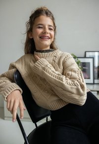 Beige kabelgebreide trui met geribde manchetten en een ronde halslijn, gecombineerd met een zwarte gestructureerde outfit, zittend op een stoel tegen een neutrale achtergrond.
