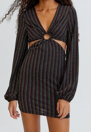 Mujer con un vestido mini negro de manga larga con rayas verticales multicolores, abertura en forma de ojo de cerradura en la parte delantera y mangas fruncidas.