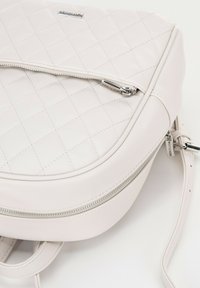 Jasno beżowy pikowany skórzany plecak z sylwerowymi zamkami błyskawicznymi i regulowanymi paskami, zaprezentowany na białym tle.