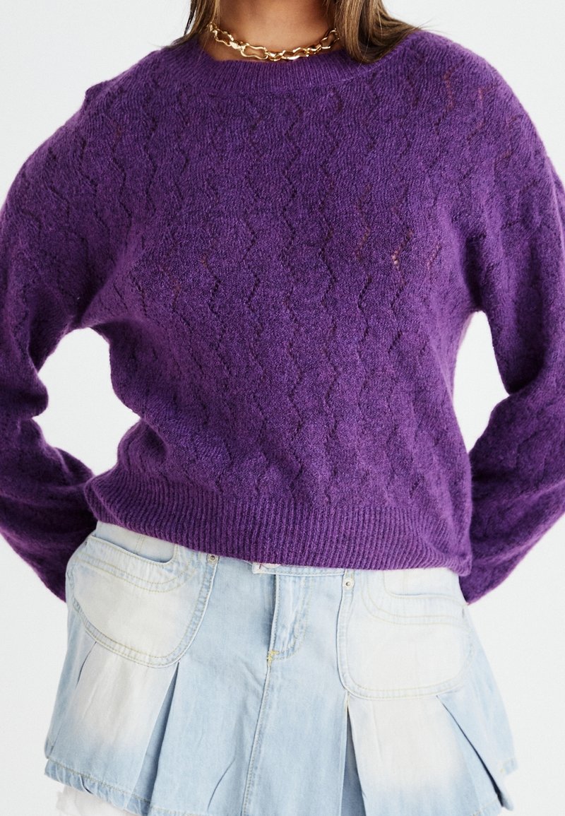 Femme portant un pull violet texturé à manches longues et une jupe en jean bleu clair avec de grandes poches avant.