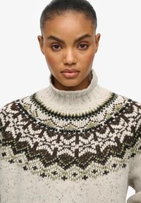 Maglione lavorato a maglia crema con colletto alto, che presenta intricate fantasie in marrone scuro, verde e blu, mettendo in mostra un design testurizzato e accogliente.