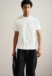 Młody mężczyzna z afro stoi trzymając czarną torbę, ubrany w prostą białą koszulkę i luźne czarne spodnie na neutralnym tle.