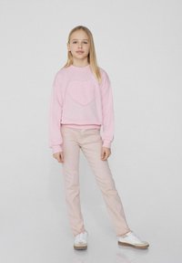 Růžová bavlněná mikina s nášivkou ve tvaru srdce, v kombinaci se světle růžovými džíny. Doplňuje ji bílá obuv se béžovými akcenty.