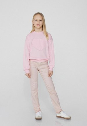 Rosa Baumwollpullover mit einem Herzpatch, kombiniert mit hellrosa Jeans. Abgerundet mit weißen Turnschuhen mit beigen Akzenten.