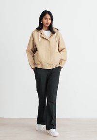 Beige hettejakke med knappelukking, elastiske mansjetter og snor, kombinert med svarte bukser og hvite sneakers, mot en ensfarget bakgrunn.