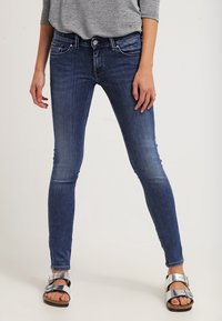 Jeans ajustados de denim azul oscuro, con una cintura de altura media, detalles de desvanecimiento sutil y un diseño clásico de cinco bolsillos. Combinados con sandalias plateadas.