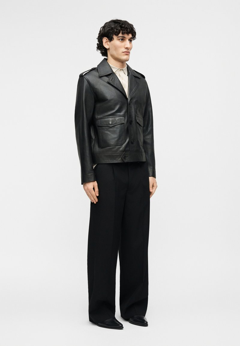 Young man wearing a black leather jacket over a beige shirt, paired with wide-leg black trousers and black shoes, standing against a white background.