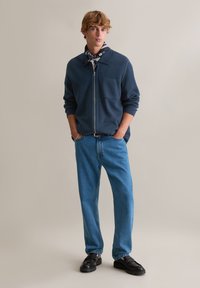 Niebieska bluza zapinana na zamek z kieszenią na piersi, zestawiona z jasnoniebieskimi dżinsami i czarnymi butami wsuwanymi. Modelka ma na sobie czarno-biały szal w wzory.