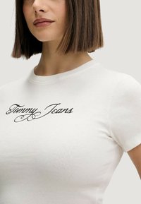 Bílé žebrované tričko s krátkými rukávy a kulatým výstřihem. Na prsou je uprostřed černé kurzívní logo "Tommy Jeans".