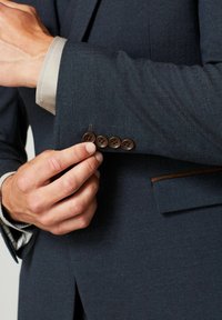 Granatowa marynarka w jodełkę, z brązowymi guzikami przy mankietach i jasnym wykończeniem kołnierzyka widocznym przy rękawie.