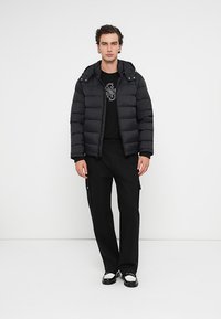 Μαύρο μπουφάν με κουκούλα, με επενδεδυμένο σχέδιο. Φορεμένο πάνω από μαύρη μπλούζα με γραφικά και μαύρο παντελόνι cargo, συμπληρωμένο με εσπαντρίγιες.