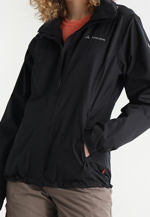 Person trägt eine schwarze Vaude Zip-Up-Jacke mit den Händen in den Taschen und beige Hosen vor einem schlichten hellen Hintergrund.
