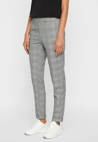 Kvinna iklädd grå rutig slim-fit-byxor med fickor, en svart topp och vita sneakers, stående mot en enfärgad bakgrund.