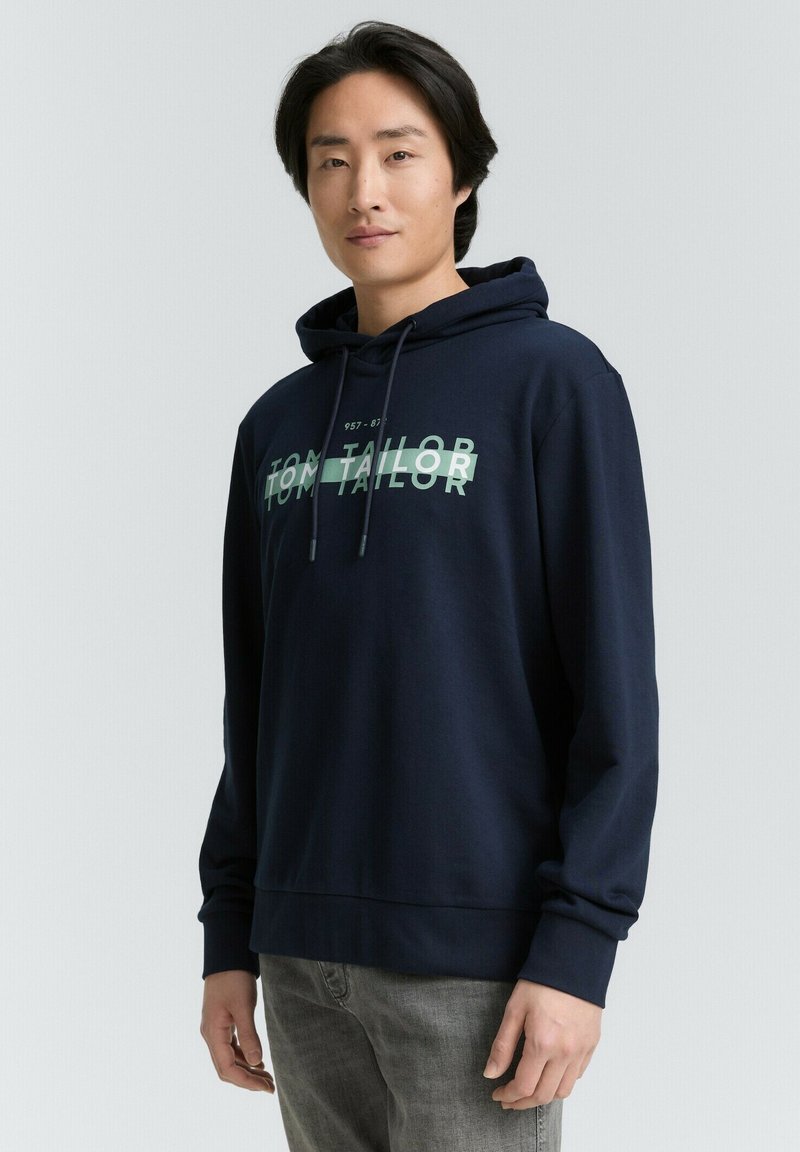 Felpa blu navy con tasca a canguro, cappuccio con coulisse e grande logo verde "TOM TAILOR" sul davanti. Tessuto morbido, maniche lunghe.